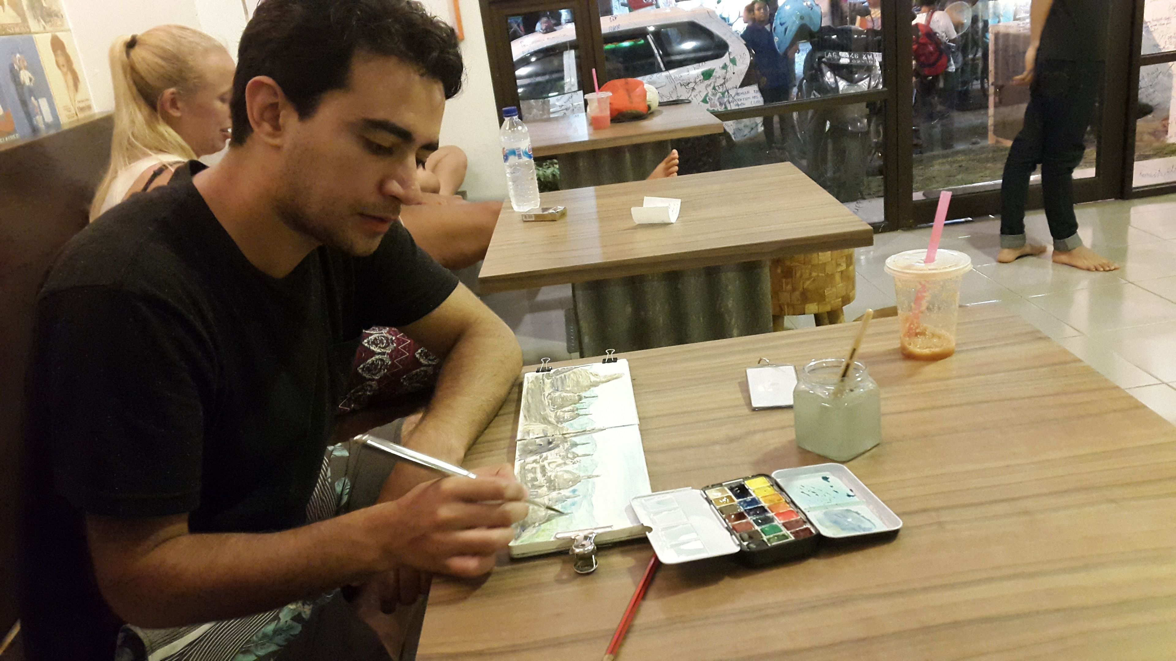 A person sitting at a table with a sketchbook, a water bottle, a drink, and a small container.
