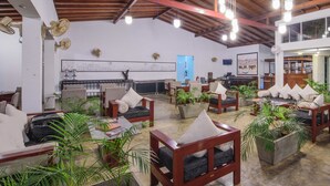 A spacious indoor area with multiple seating arrangements, including sofas and chairs, surrounded by potted plants. There are ceiling fans and a visible fire extinguisher on the wall. - Hotel Wewa Addara (Sigiriya)