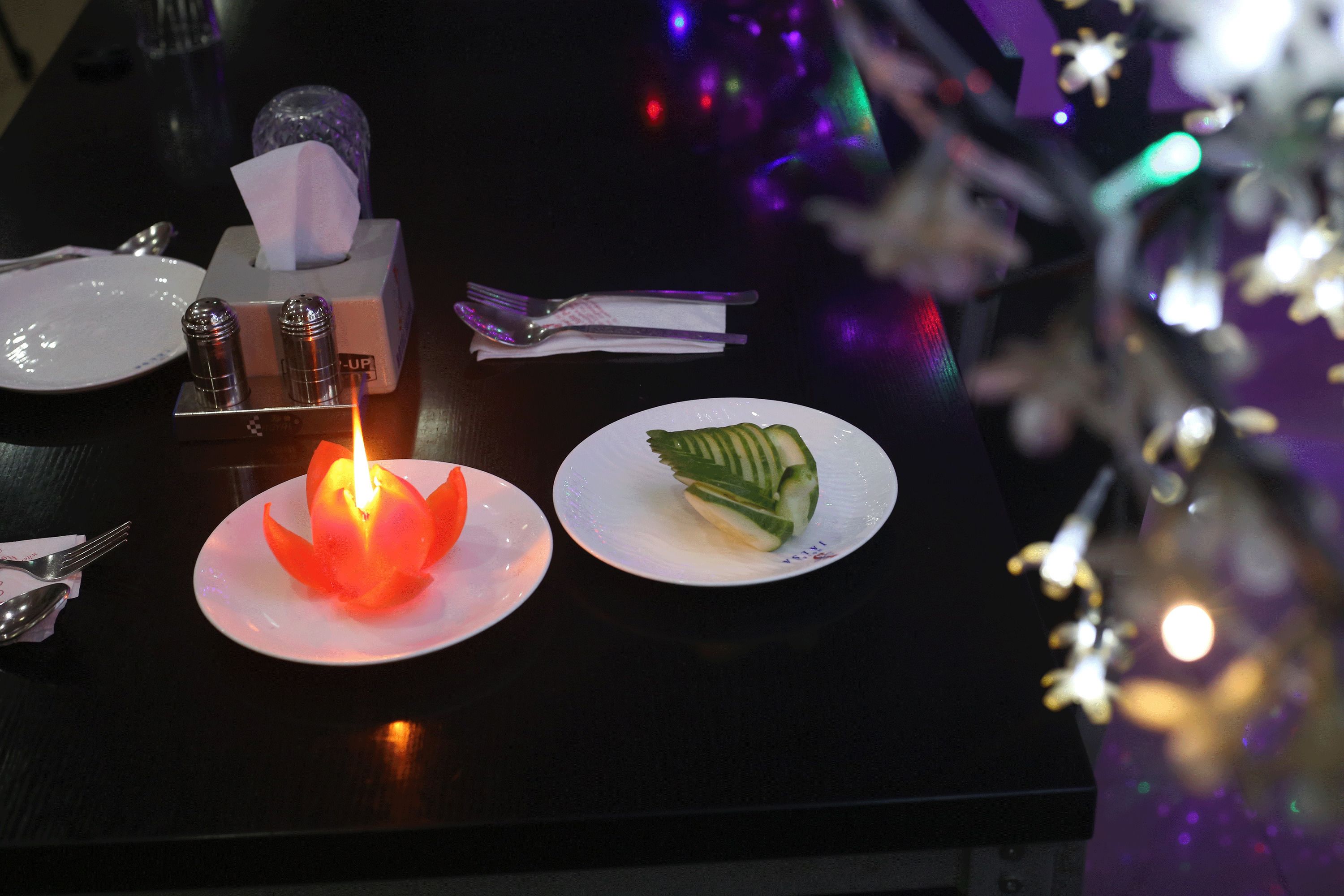 Una mesa con una vela encendida, un caja de pañuelos, un tenedor, una cuchara y un plato con una rodaja de pepino.
