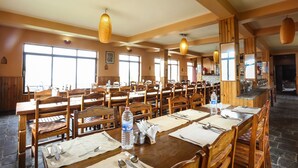 A spacious dining area with multiple wooden tables and chairs, large windows allowing natural light, and hanging woven light fixtures. - Dhaulagiri View Hotel (Pokhara)