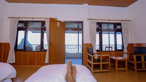 A hotel room with a bed, a desk, and a chair. There are curtains on the windows, and a balcony is visible through the open door. - Dhaulagiri View Hotel (Pokhara)