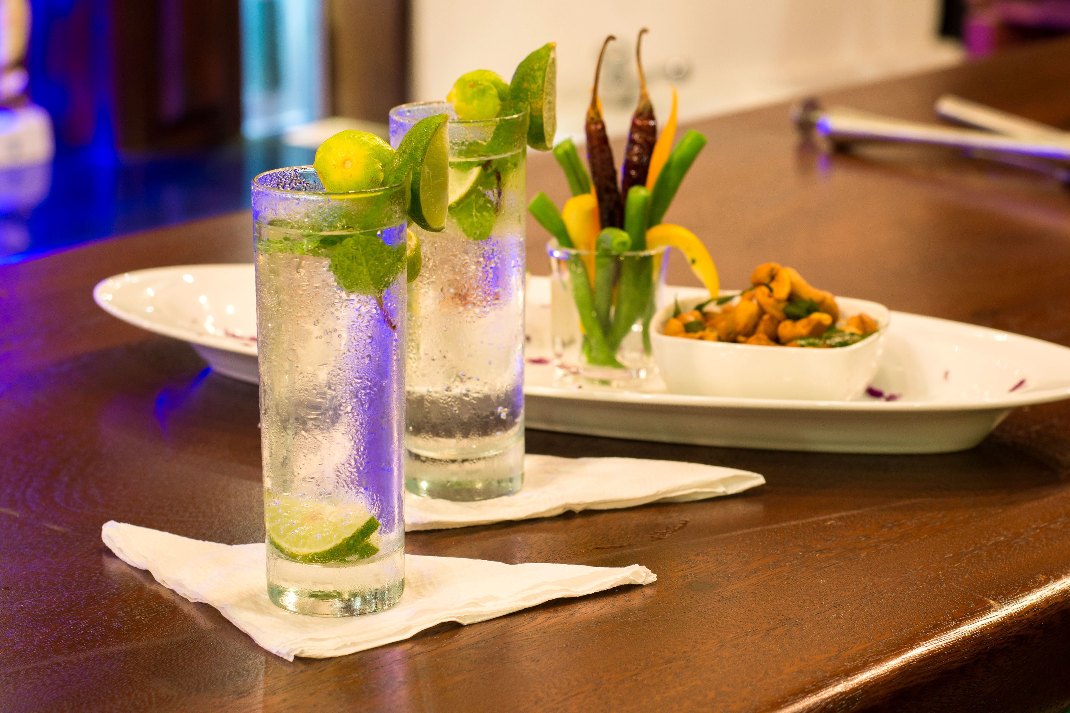 Two glasses of a lime and mint beverage on a wooden table with a plate of nuts and a small bowl of herbs in the background.