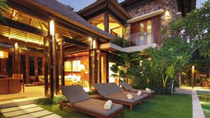 A well-lit outdoor lounge area with wooden loungers and a stone building in the background. - Villa Suar Drupadi (Seminyak)