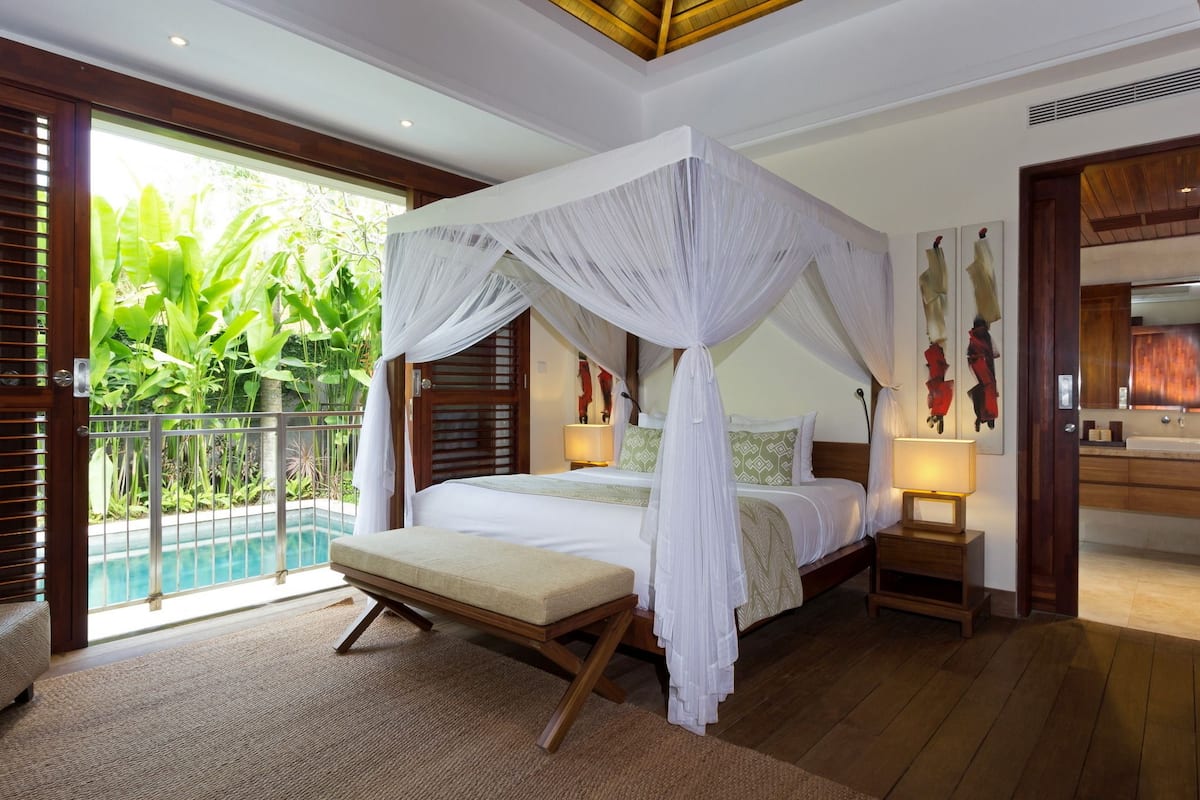 A bedroom with a four-poster bed, a bench, a bedside table, and a view of a pool.