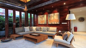 A modern living room with a sectional sofa, a coffee table, and a side chair. There are large windows, a wooden ceiling, and a lamp. - Villa Suar Drupadi (Seminyak)