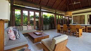 A spacious living area with a wooden ceiling, a sofa set, a dining table with chairs, and a kitchen area visible through large glass doors. - Villa Suar Drupadi (Seminyak)