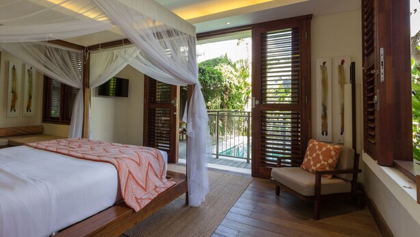 Quarto com cama, uma cadeira, varanda com persianas de madeira e vista para áreas verdes.