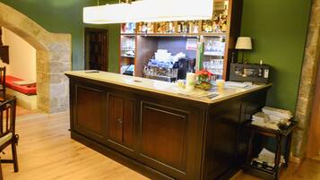 A bar area with a wooden counter, shelves with bottles, a coffee machine, and a small table with a lamp.