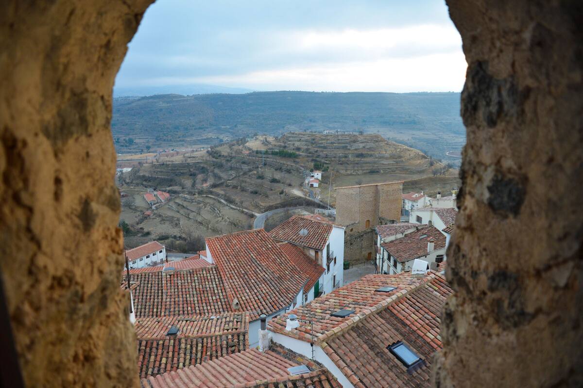 Blick auf Gebäude mit Terrakotta-Dächern durch einen Steinbogen, mit Blick auf eine hügelige Landschaft.