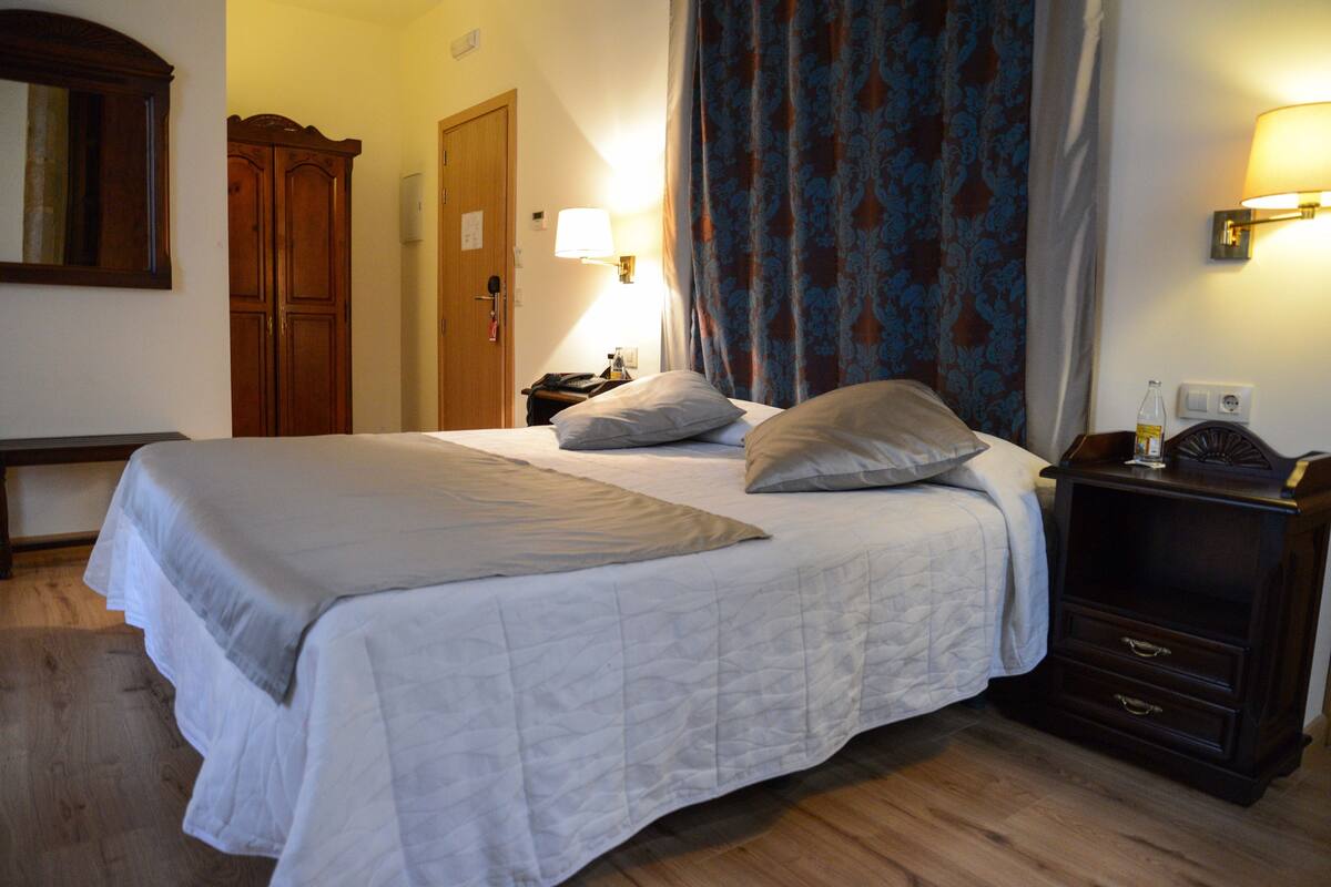 Ein ordentlich bezogenes Bett mit weißer Bettdecke und Kissen, ein Holzschrank, ein Nachttisch mit Lampe und ein gemustertes Vorhang.