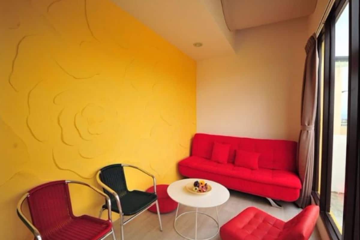 A room with a red sofa, a red chair, a black chair, and a small white table with a bowl of fruit.
