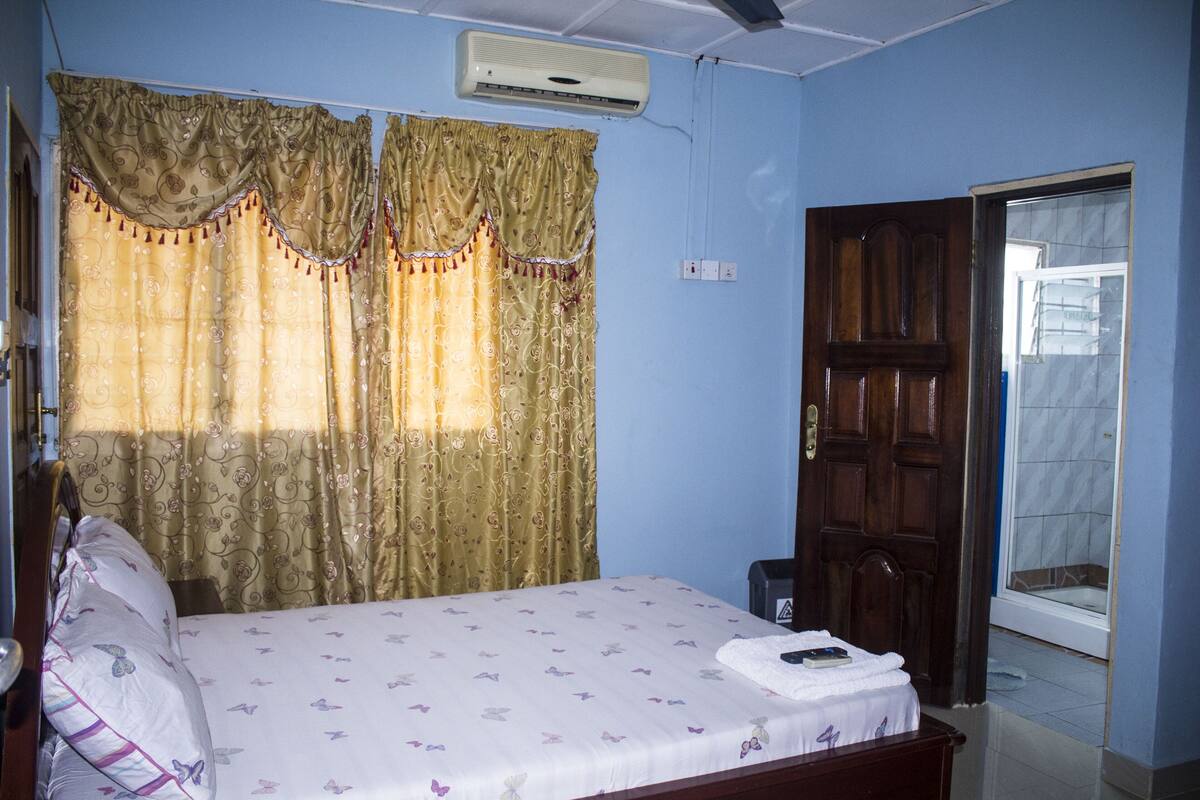 A bedroom with a bed, curtains, an air conditioner, a wooden door, and a bathroom visible through the door.