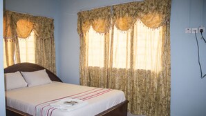 A neatly made bed with a white bedspread and a red and white striped blanket, a wooden headboard, and a window with patterned curtains. - Adinkra Lodge (Accra)