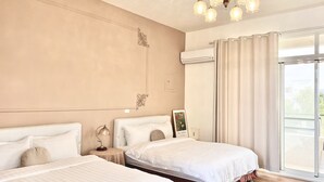 Habitación de hotel con dos camas, un candelabro, un equipo de aire acondicionado y un ventanal con cortinas.