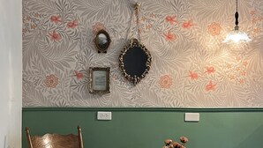 Una habitación con papel tapiz floral, una mecedora, una mesita con un jarrón de flores y una lámpara.