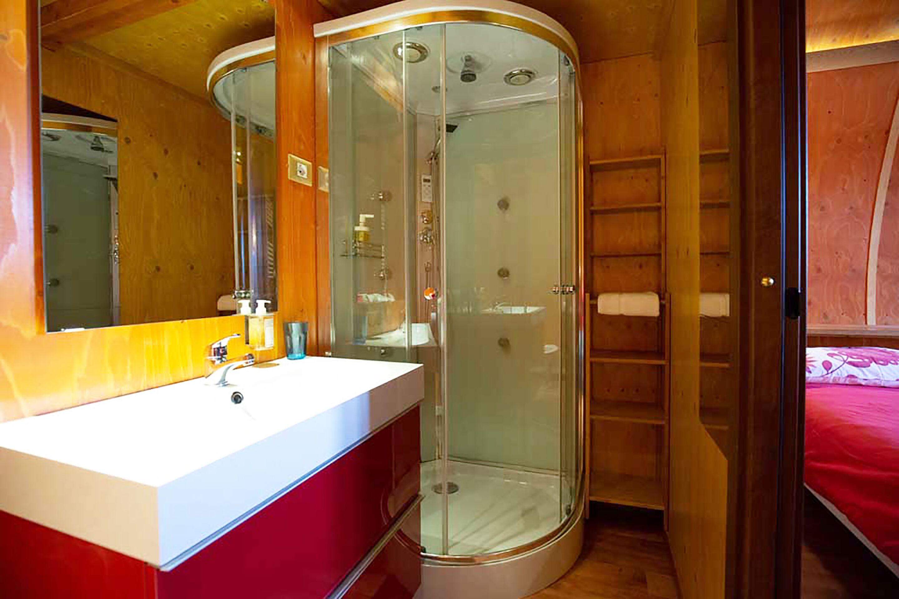 Een badkamer met een douchecabine van glas, een witte wastafel met een rode kast, en een houten inrichting.