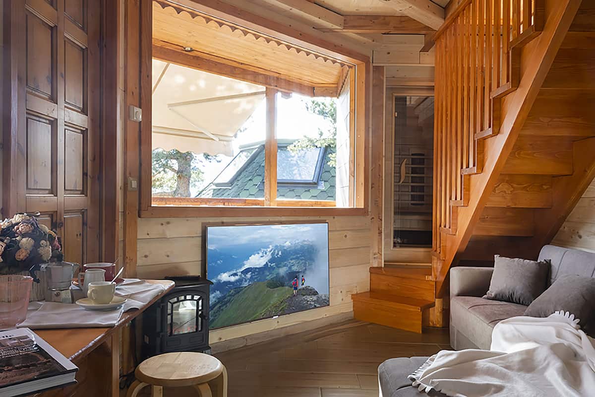 Interior acogedor de una cabaña con una escalera de madera, un televisor montado en la pared y una zona de comedor con mesa y sillas.