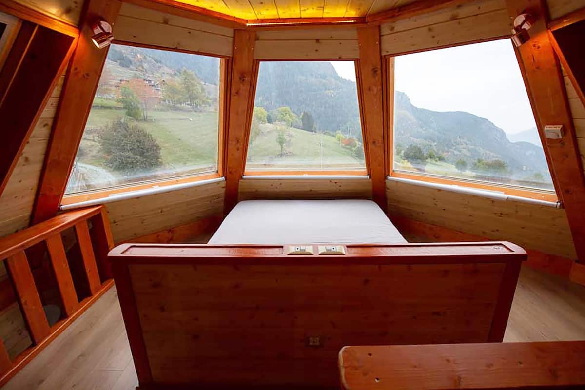 Una cabaña de madera con cama y ventanas amplias que ofrecen vistas a un paisaje montañoso.