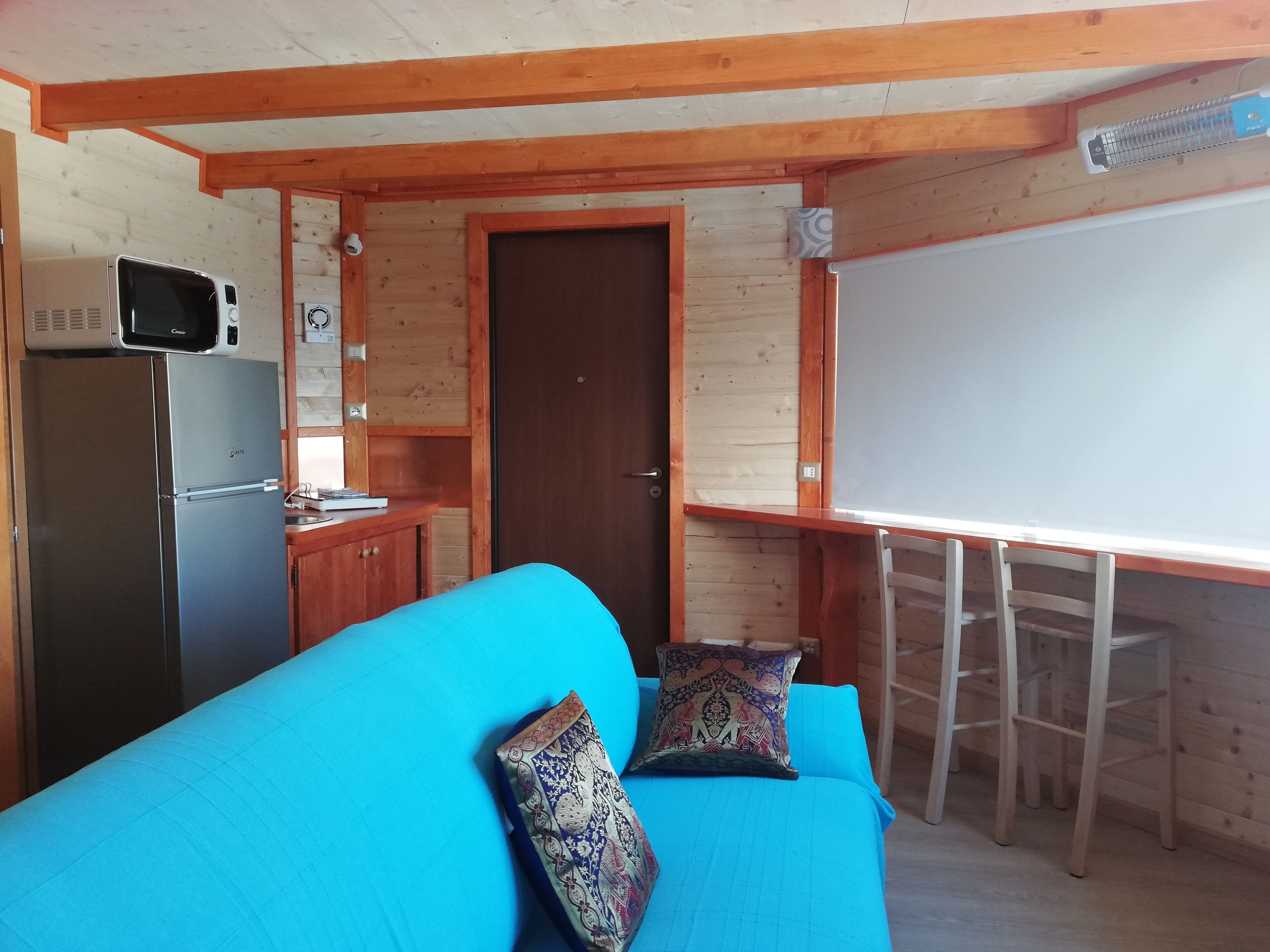 Una cocina compacta con microondas, refrigerador y gabinetes de madera. Un sofá azul con cojines estampados está ubicado frente a una pizarra y una mesa de madera.