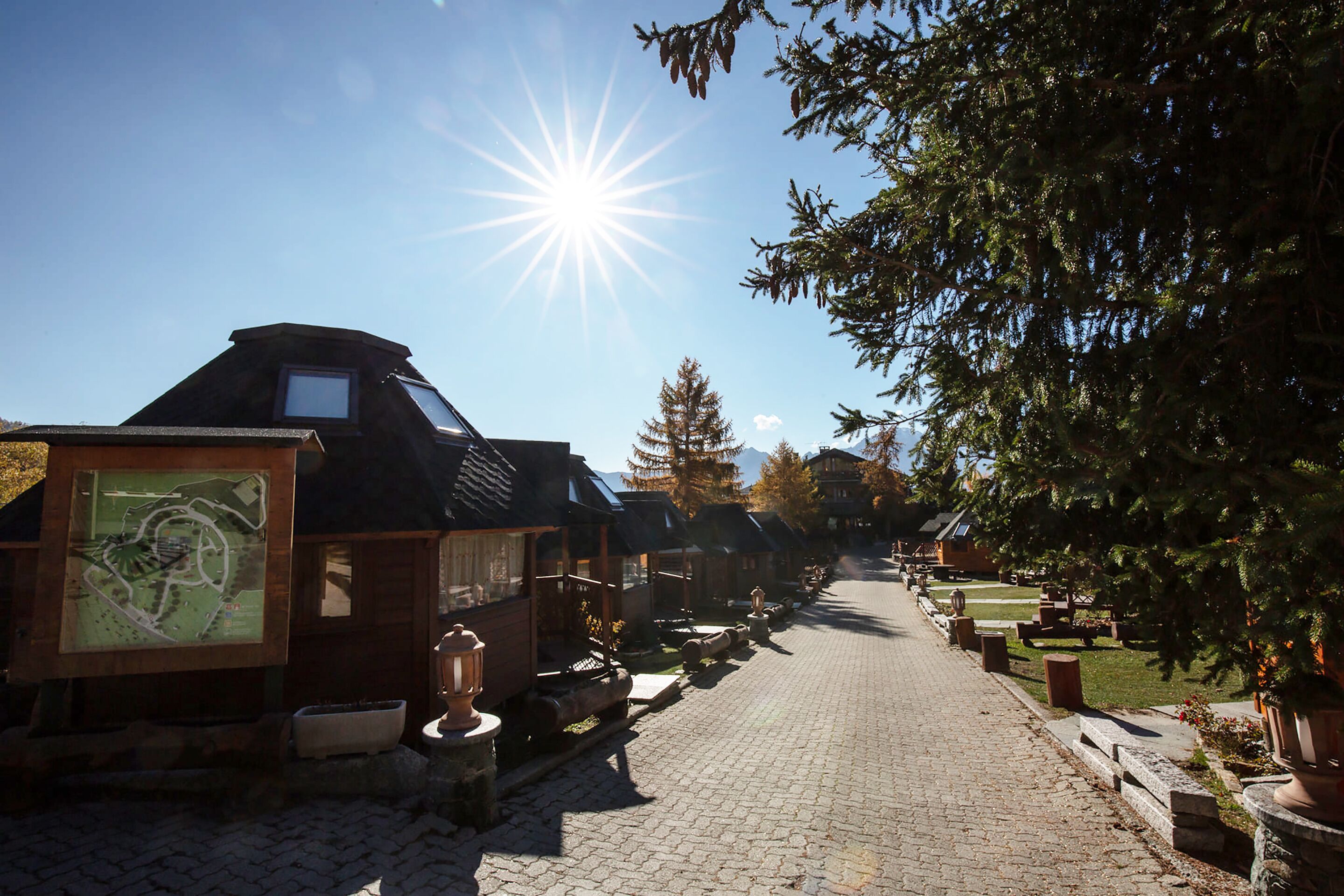 A cobblestone street lined with wooden buildings, some with sloped roofs and large windows, under a clear sky with the sun shining brightly.
