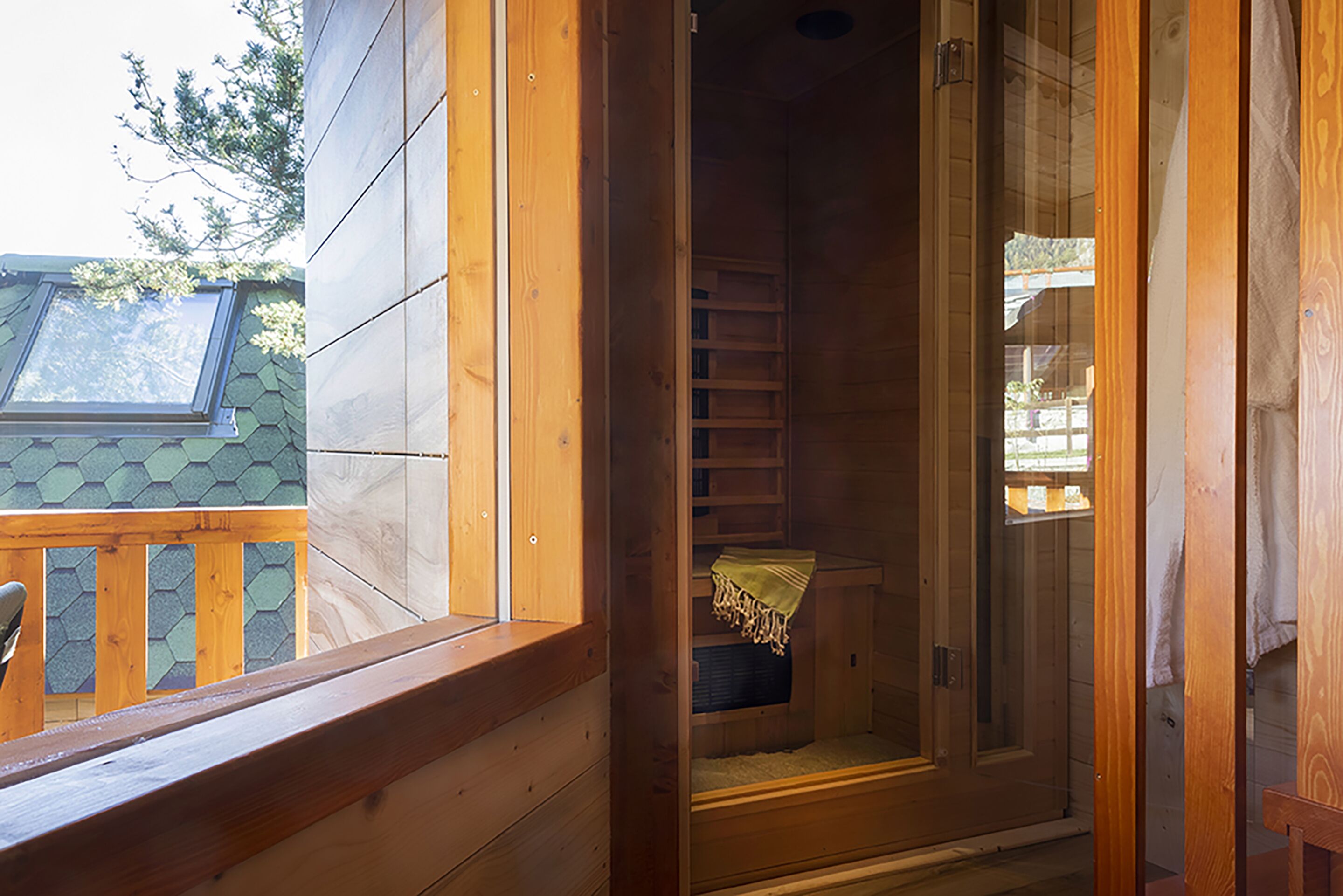 Une cabane en bois avec une porte en verre donnant sur une petite salle de bain fermée, équipée d’un banc et d’une serviette accrochée au mur.