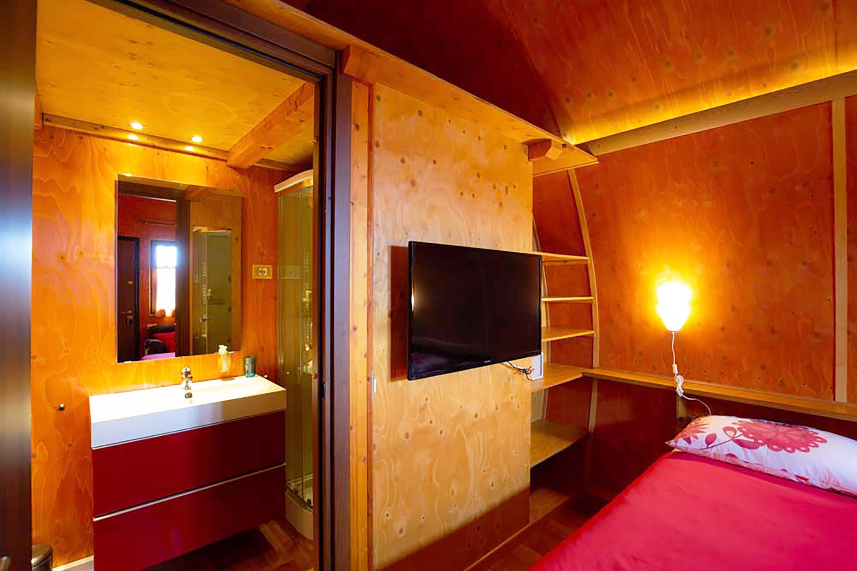 Ein Zimmer in einer Holzhütte mit Holzwänden, einem an der Wand montierten Fernseher, einem Bett mit roter Decke, einer Holzleiter und einem Badezimmer mit Waschbecken und Spiegel.