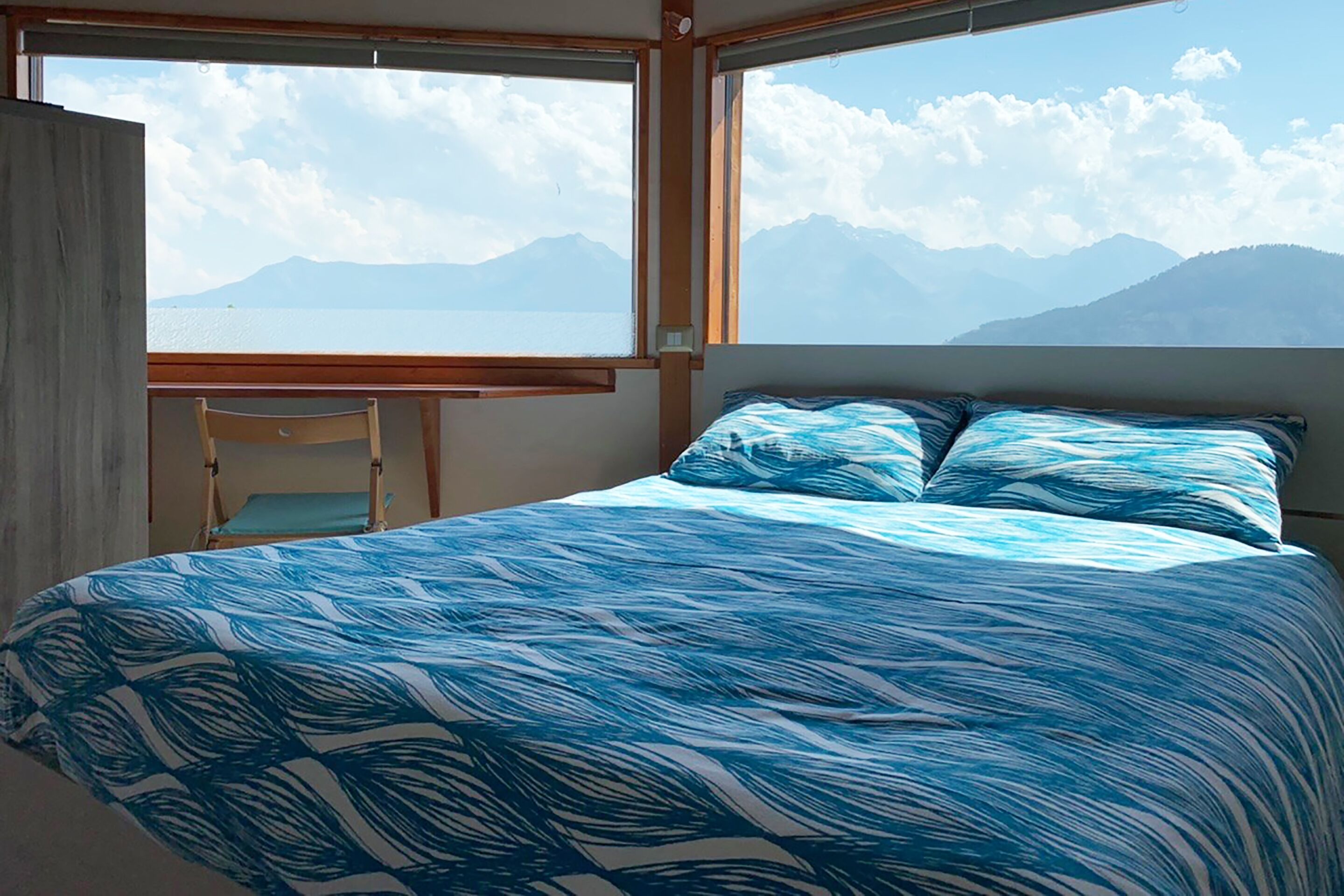 A bedroom with a bed, a chair, and a table. It has a view of mountains and a body of water.