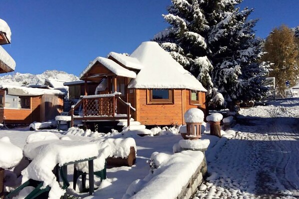 A wooden cabin with a snow-covered roof and surrounding snow, set in a mountainous area.