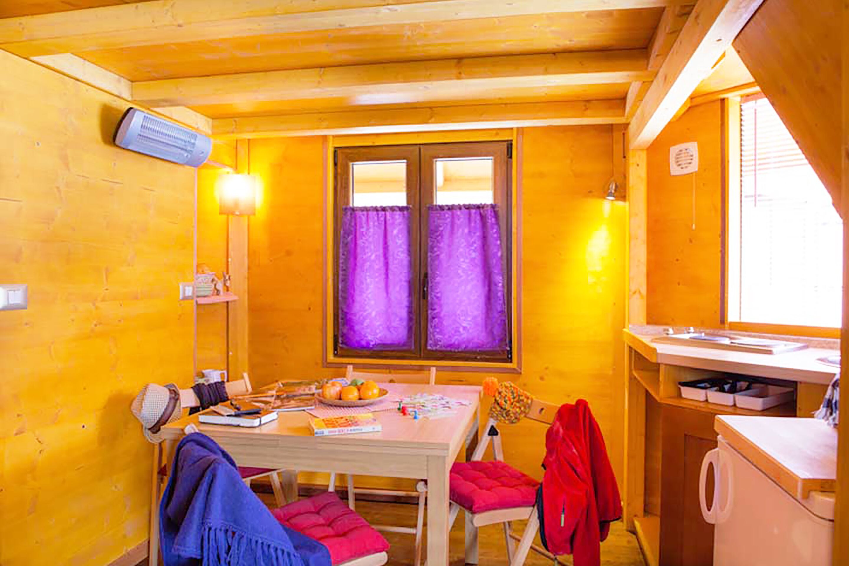 Una habitación acogedora con detalles de madera, una mesa de comedor con sillas, una ventana con cortinas moradas y una zona de cocina con un fregadero y armarios.