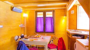 Una habitación acogedora con detalles de madera, una mesa de comedor con sillas, una ventana con cortinas moradas y una zona de cocina con un fregadero y armarios.
