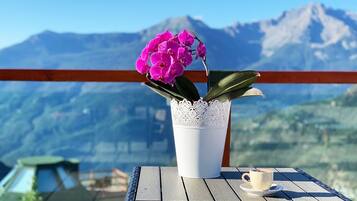 A white vase with pink flowers on a table with a cup of coffee, overlooking a mountainous landscape.