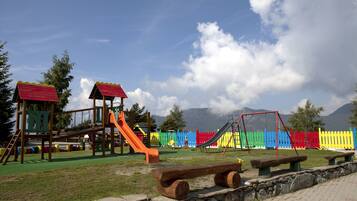 Children can play on the slide and swings in this playground surrounded by a fence and set against a mountain backdrop.