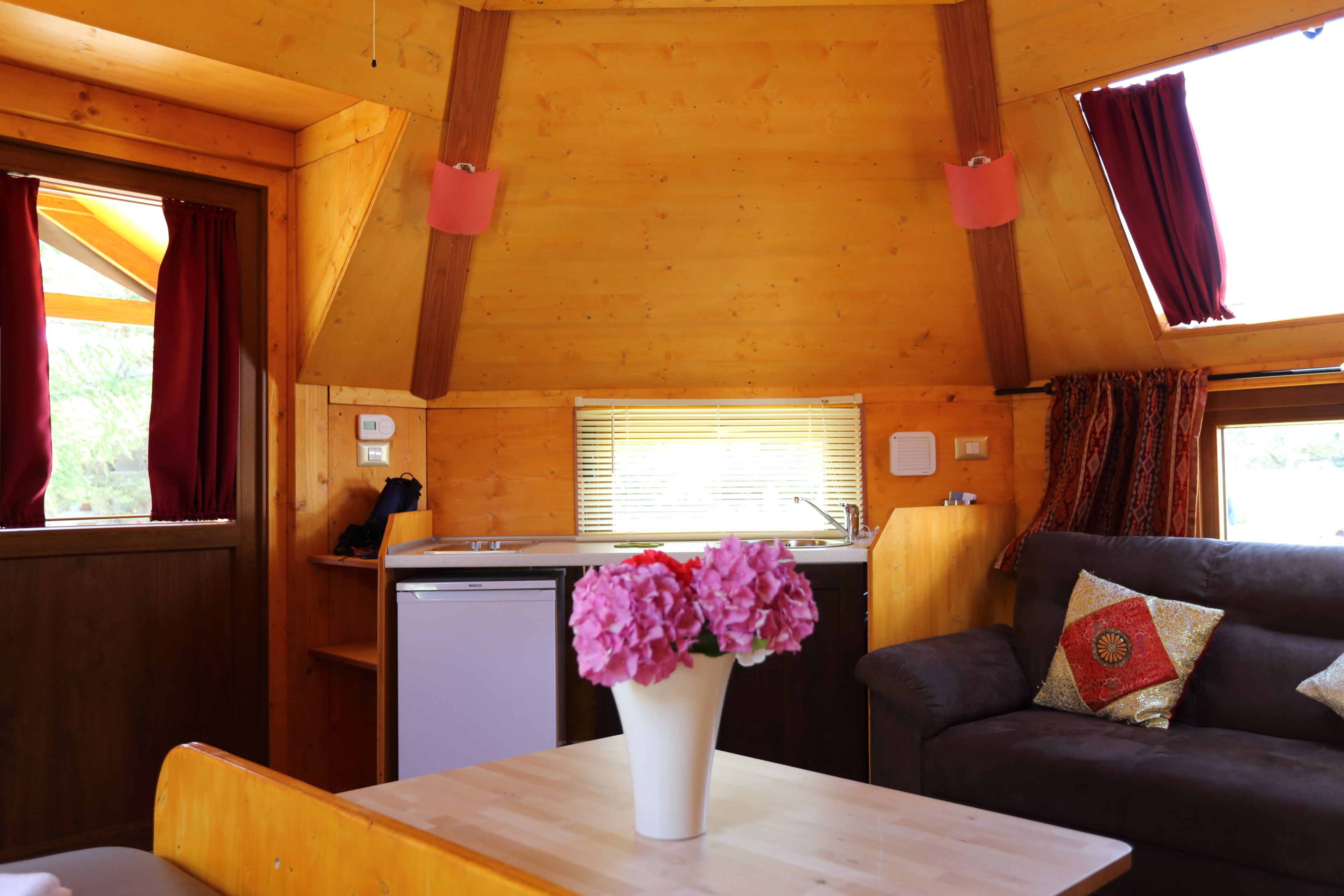 A cozy cabin interior with a wooden ceiling, a dining table with a vase of flowers, a sofa with decorative pillows, and a kitchen area with a dishwasher.