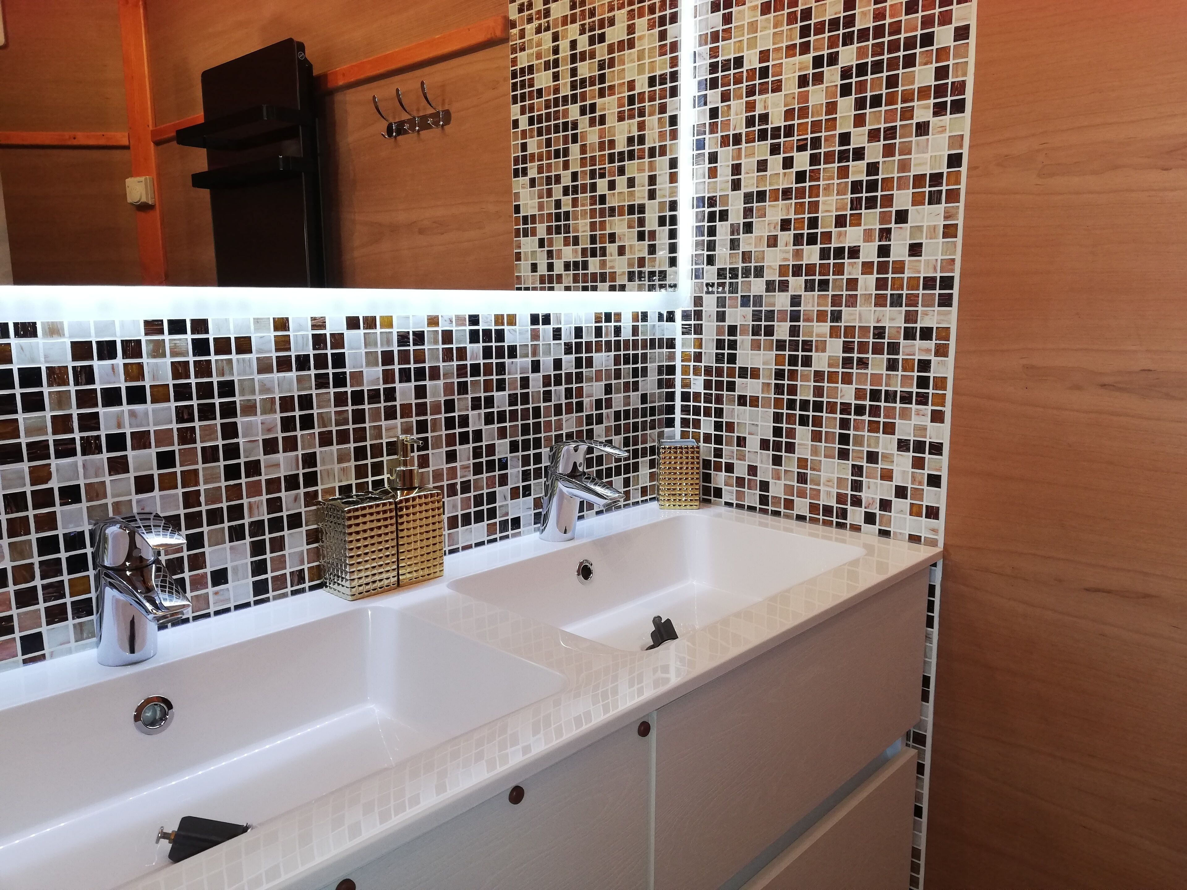 Un baño con lavamanos blanco, dos grifos y un espejo. La pared está revestida con un mosaico.