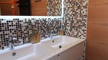 Un baño con lavamanos blanco, dos grifos y un espejo. La pared está revestida con un mosaico.