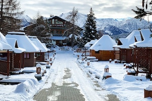 Un sendero cubierto de nieve que lleva a un albergue de madera con varias cabañas, rodeado de montañas nevadas.
