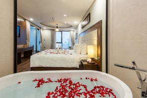 Ein Hotelzimmer mit einem großen Bett, einem Badezimmer mit einer Badewanne voller Rosenblätter und Blick auf die Stadt durch große Fenster.