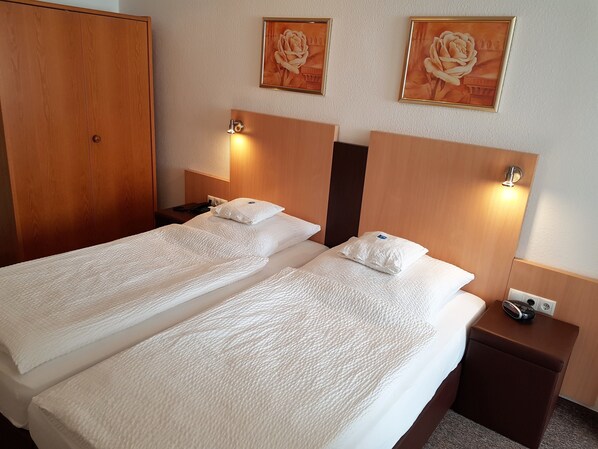 A hotel room with two single beds, wooden headboards, bedside tables, a nightstand, and two framed floral paintings on the wall. - Hotel zum Anker (Andernach)
