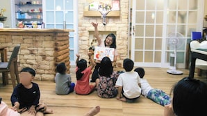 Un grupo de niños sentados en el suelo de una habitación con pared de piedra, mostrador de madera y un ventilador al fondo.