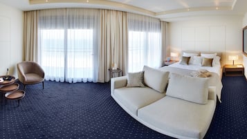 A hotel room with a curved sofa, a bed with multiple pillows, a small table with a lamp, and a chair.