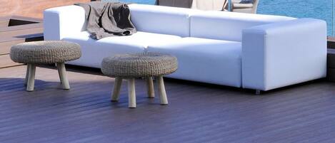 A modern outdoor seating area with a white sofa, two woven ottomans, and a small table with a bottle and glasses.