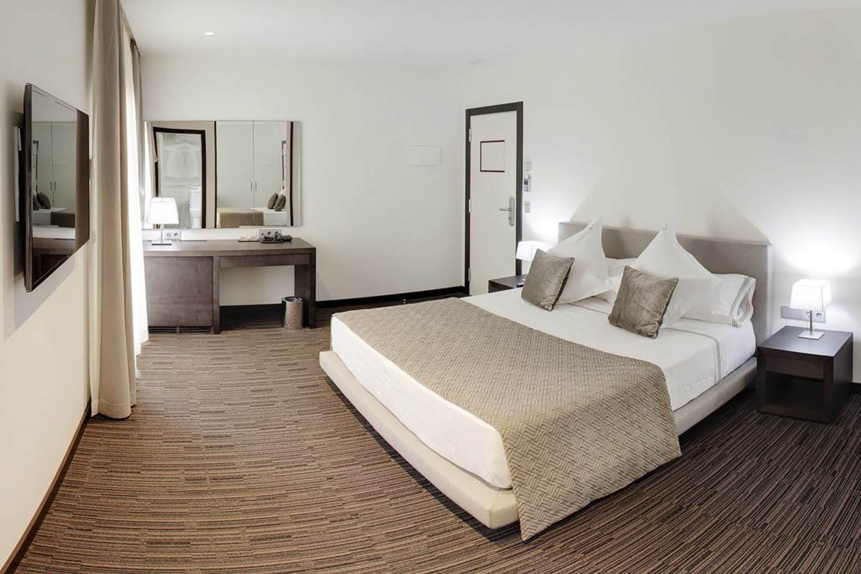 Una habitación de hotel con cama, escritorio, silla, espejo y mesita de noche.