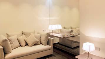A modern hotel room with a beige sofa, two bedside tables with lamps, and a small side table with a lamp.