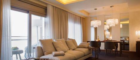 A modern hotel room with a large sofa, dining table, and chairs. There is a large window with curtains, a chandelier, and a mirror on the wall.