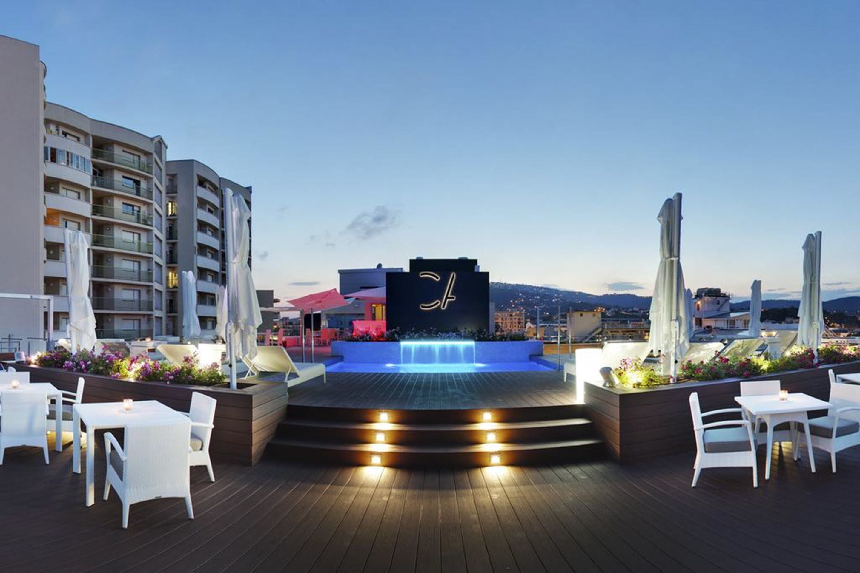 A rooftop terrace with a water feature, white outdoor seating, and a view of the cityscape.