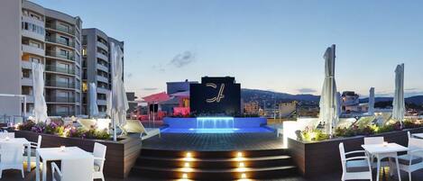 Une terrasse sur le toit agrémentée d’un élément aquatique, de mobilier d’extérieur blanc et offrant une vue sur le paysage urbain.