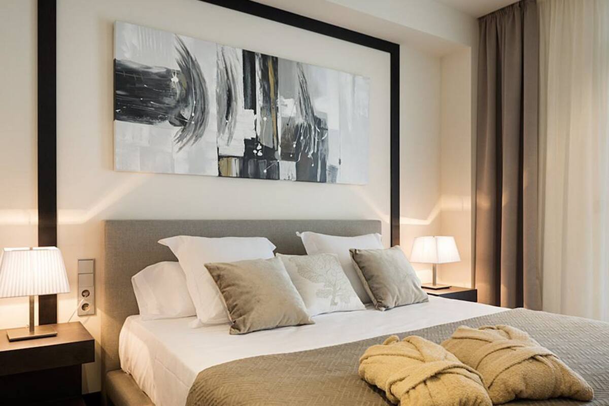 Hotelkamer met een groot bed, meerdere kussens, twee nachtkastjes met lampen, een groot abstract schilderij aan de muur en een raam met gordijnen.