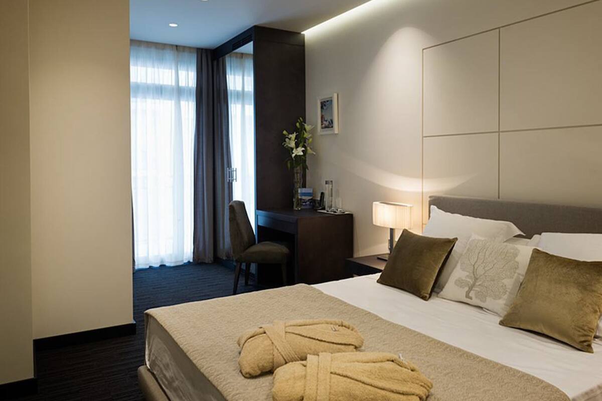 Een hotelkamer met een groot bed, twee kussens, een nachtkastje met een lamp, een stoel en een bureau met een bloemenvaas.