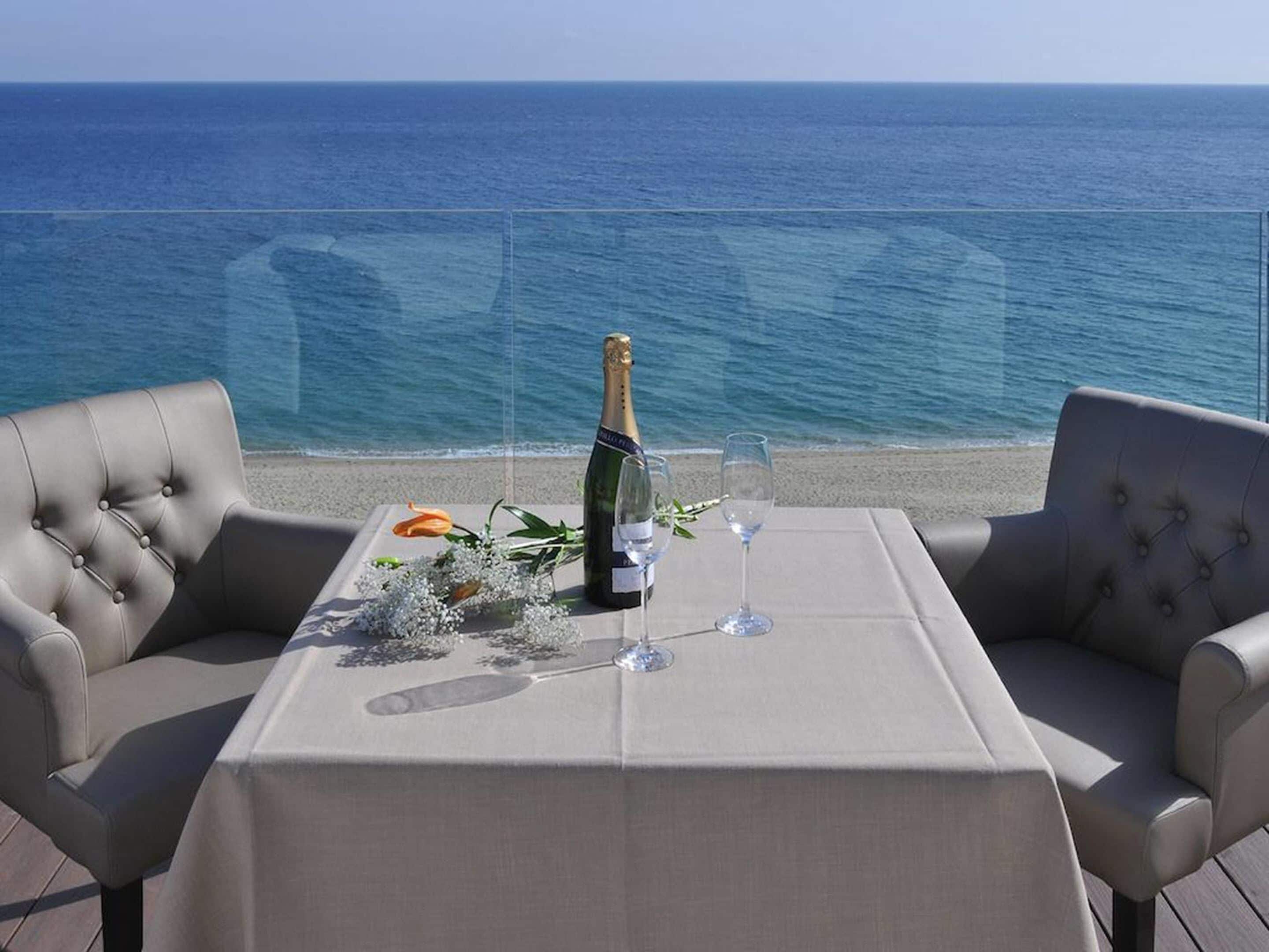 Mesa para dos con una botella de champán, copas y un ramo de flores en una terraza con vistas al mar.