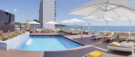 Uma área de piscina no terraço com espreguiçadeiras e guarda-sóis, vista desobstruída para o mar e um edifício alto ao fundo.